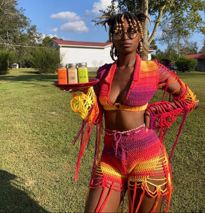 Person in colorful crochet outfit holding a tray with colorful containers outdoors.